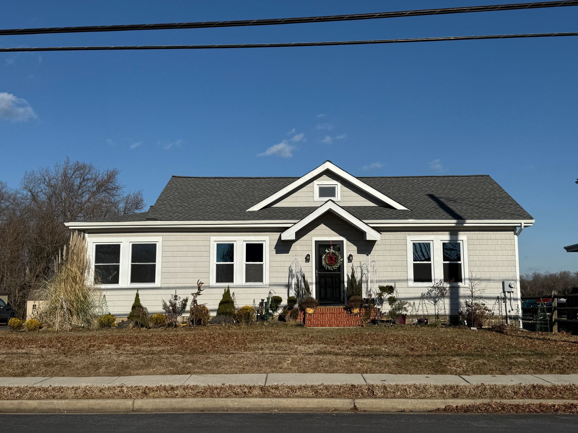 Tan house with white trim, front porch, and a dark wreath on the door. Brown grass and clear blue sky.