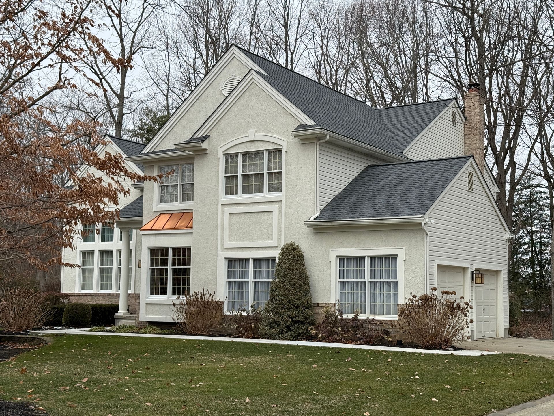 Two-story beige house with dark roof, copper-colored accent, white-framed windows, and a small, evergreen bush.