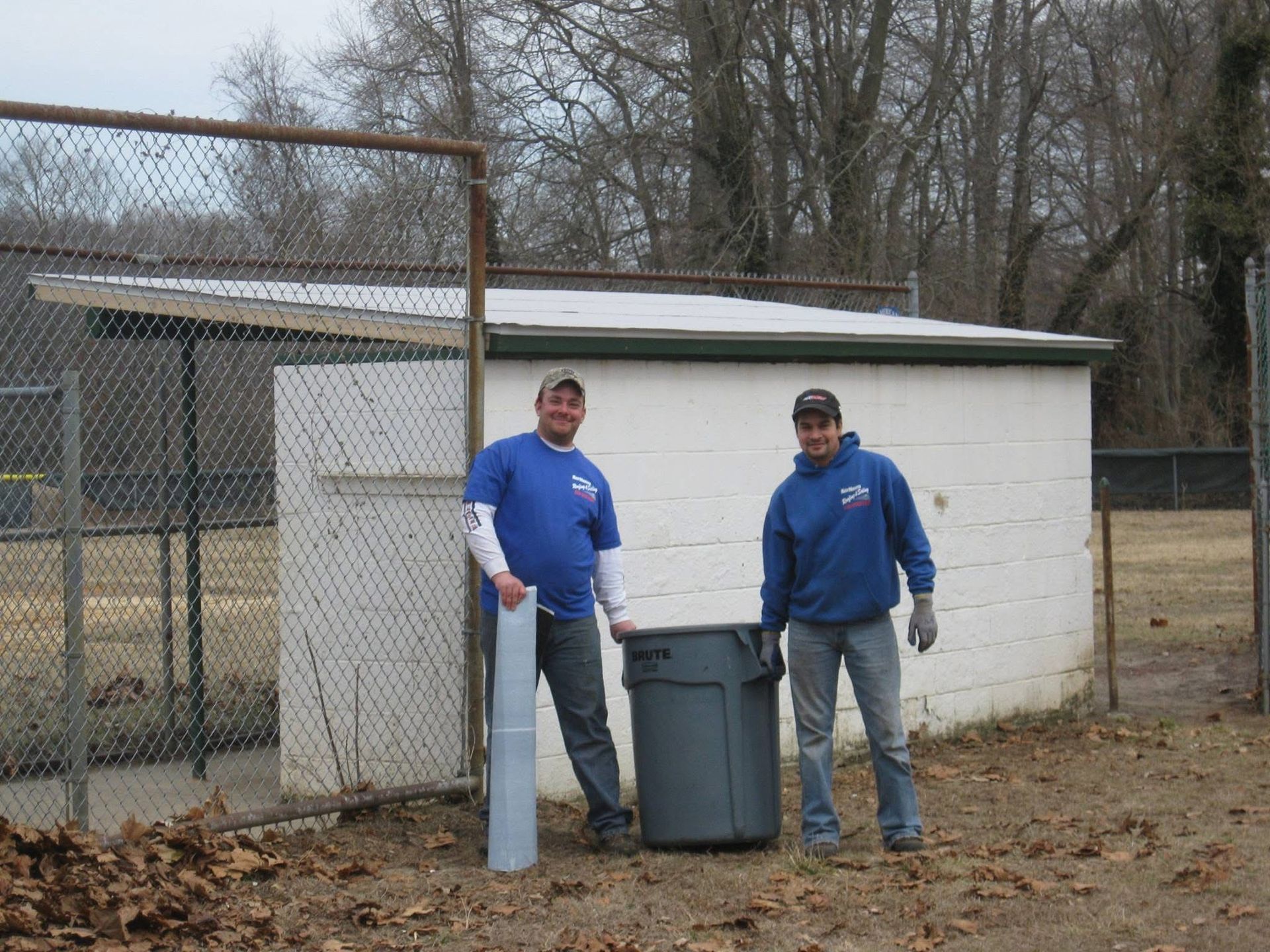 Two men in blue shirts stand outside a white building, holding trash can and rolled material; outdoor setting.