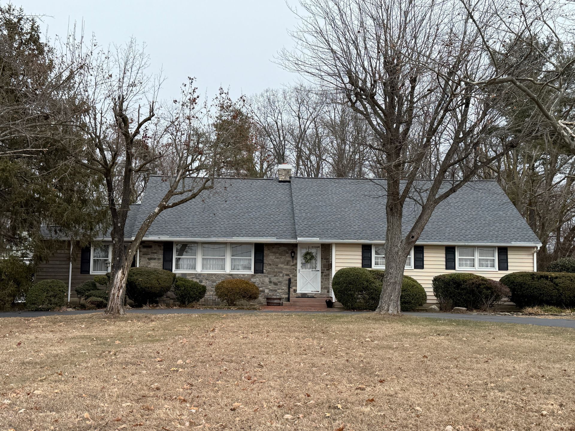 Ranch-style house with stone and beige siding, dark roof, and bare trees in the front yard. Overcast sky.