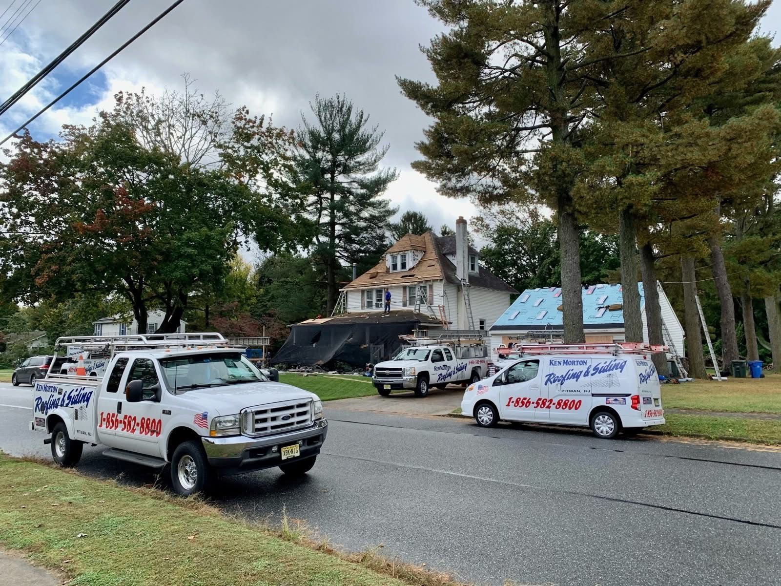 Roofing trucks parked in front of a house under construction, New Jersey, with trees on a cloudy day.