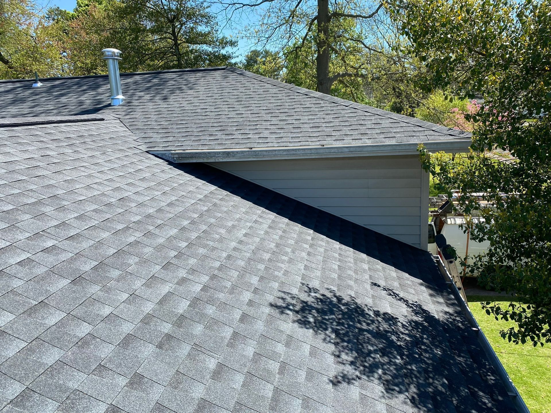 Gray asphalt shingle roof of a house, with a chimney and surrounding trees in the background.
