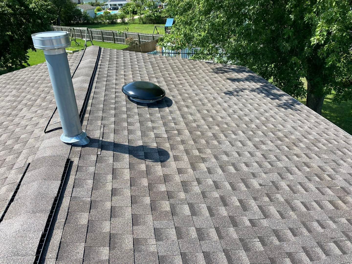 Gray shingled roof with a vent and chimney. Sunny day with green trees in the background.