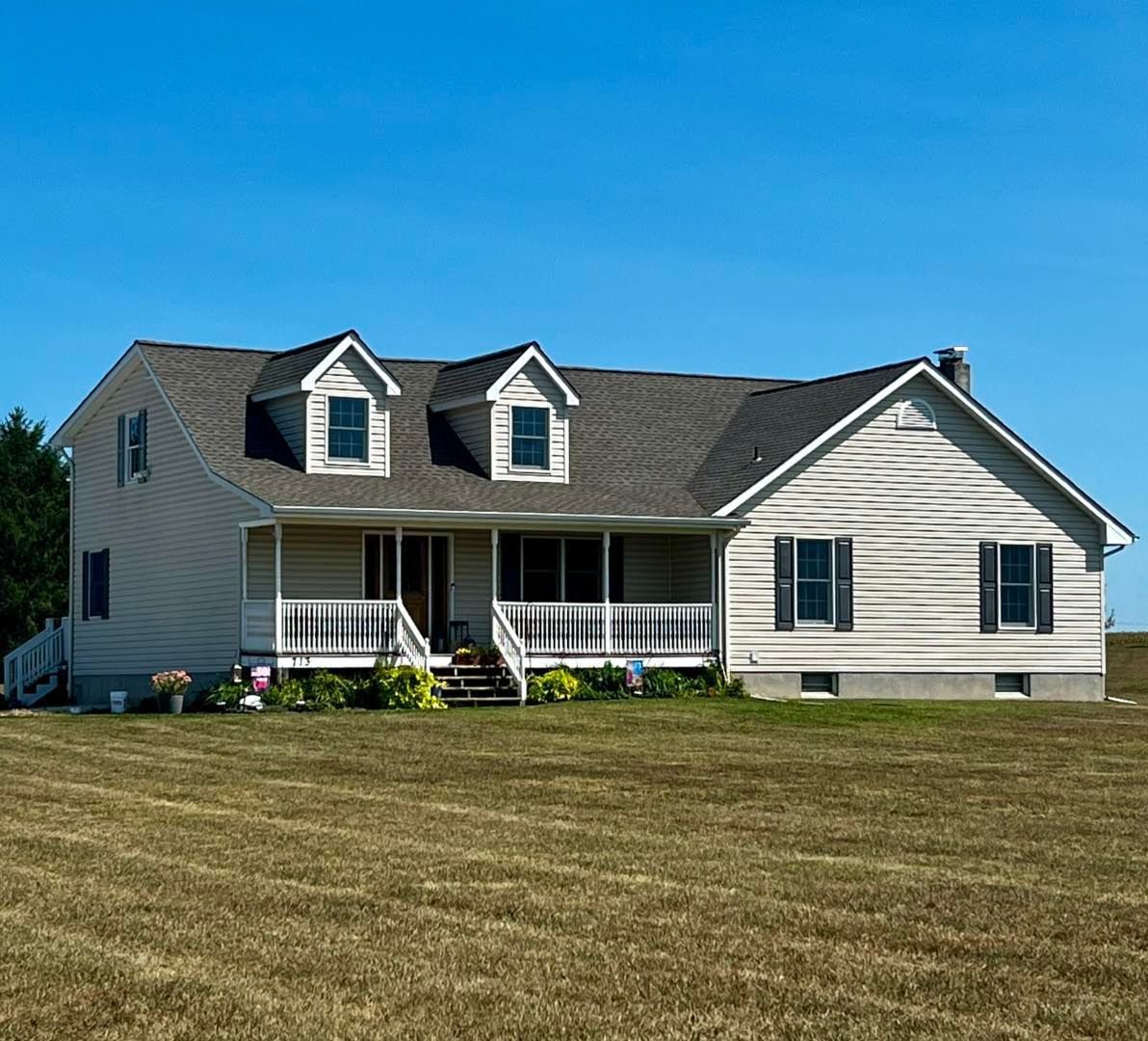 Tan two-story house with a porch, dormer windows, and gray roof on a grassy lot under a blue sky.