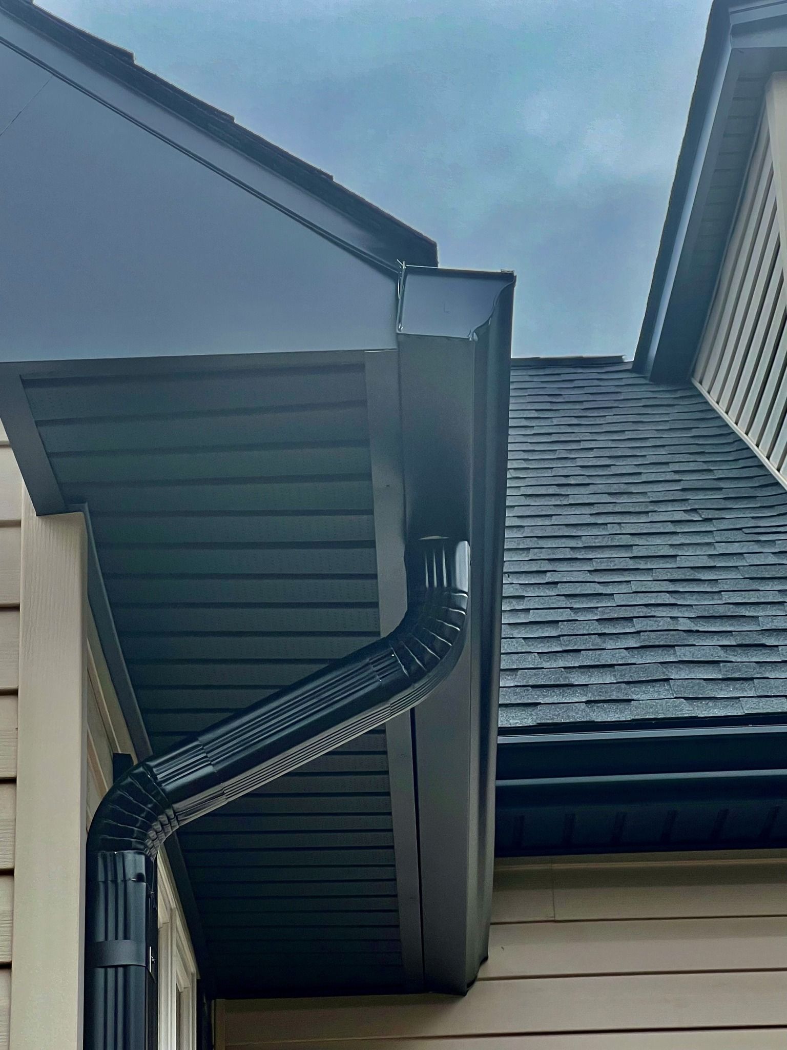 Black gutters and downspout on a house with a dark roof, against a cloudy sky.