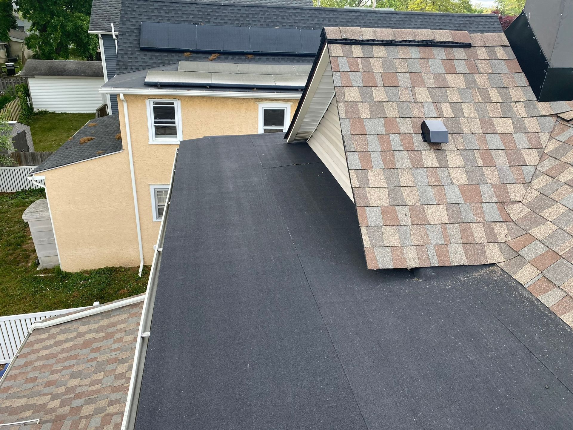 Overhead view of multiple roofs: a black flat roof next to a brown shingle roof, with a yellow house in the background.