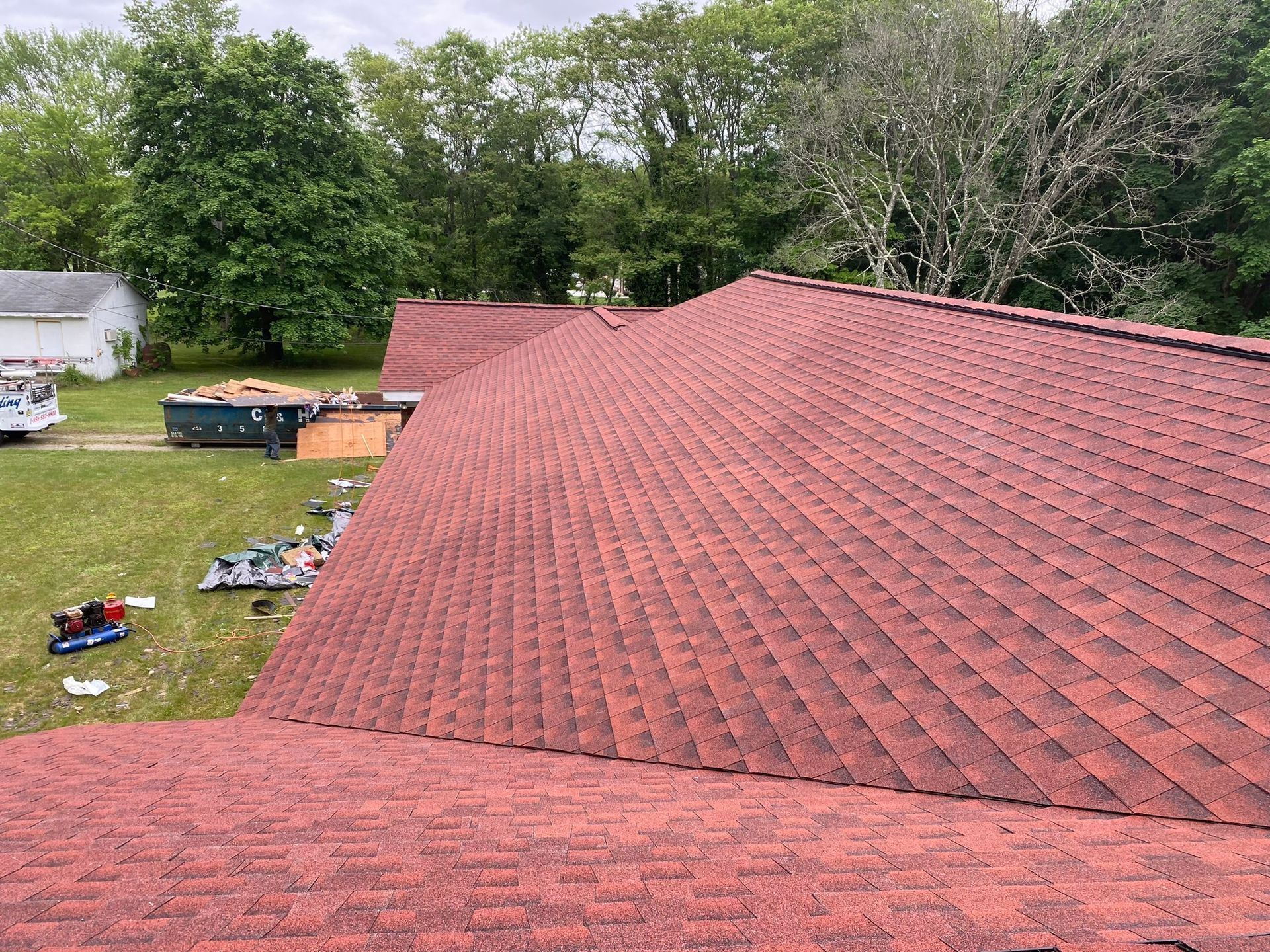 Red asphalt shingle roof on a house, trees and grass in background.