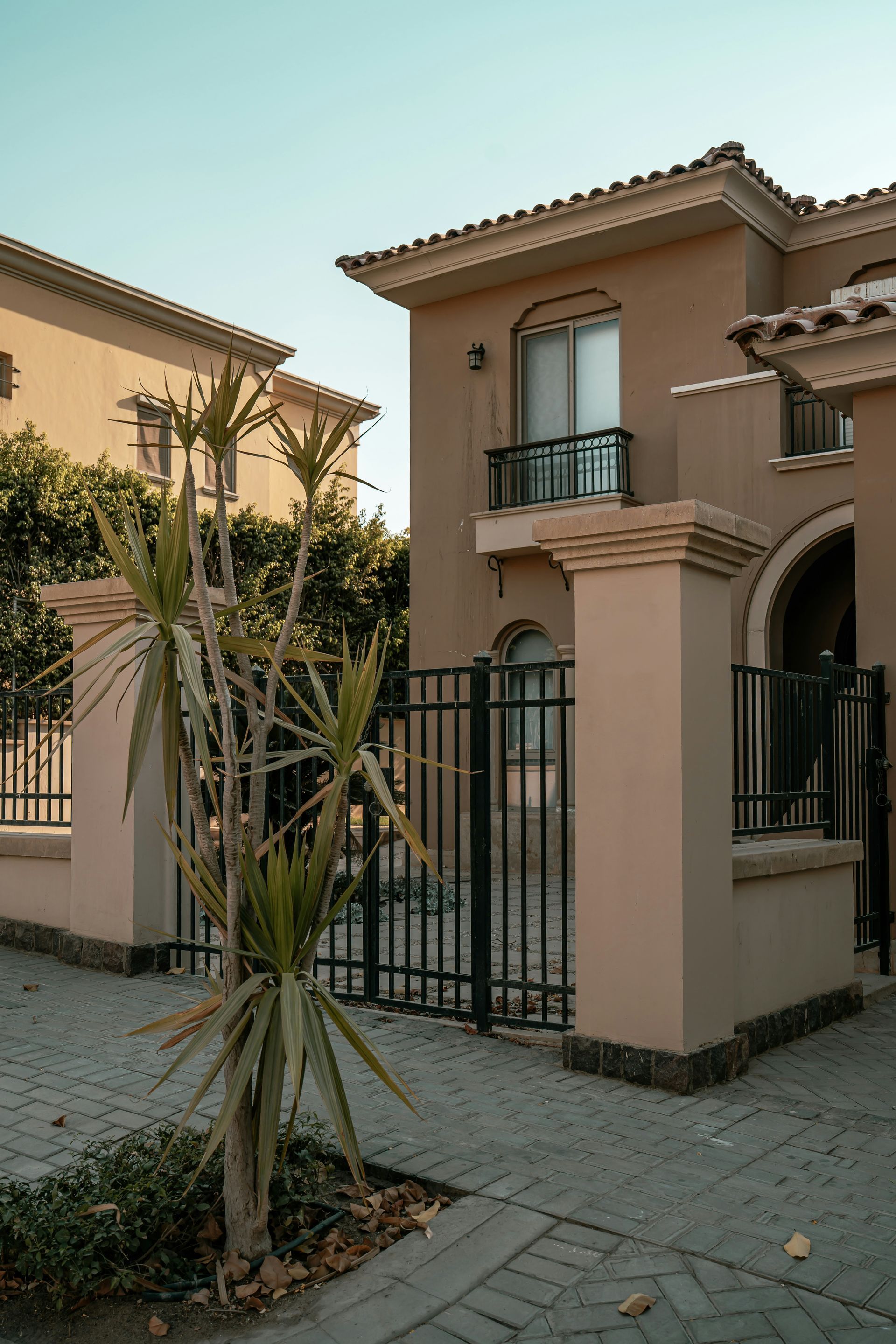 Two-story beige house with black gates and a small palm tree in a paved courtyard.