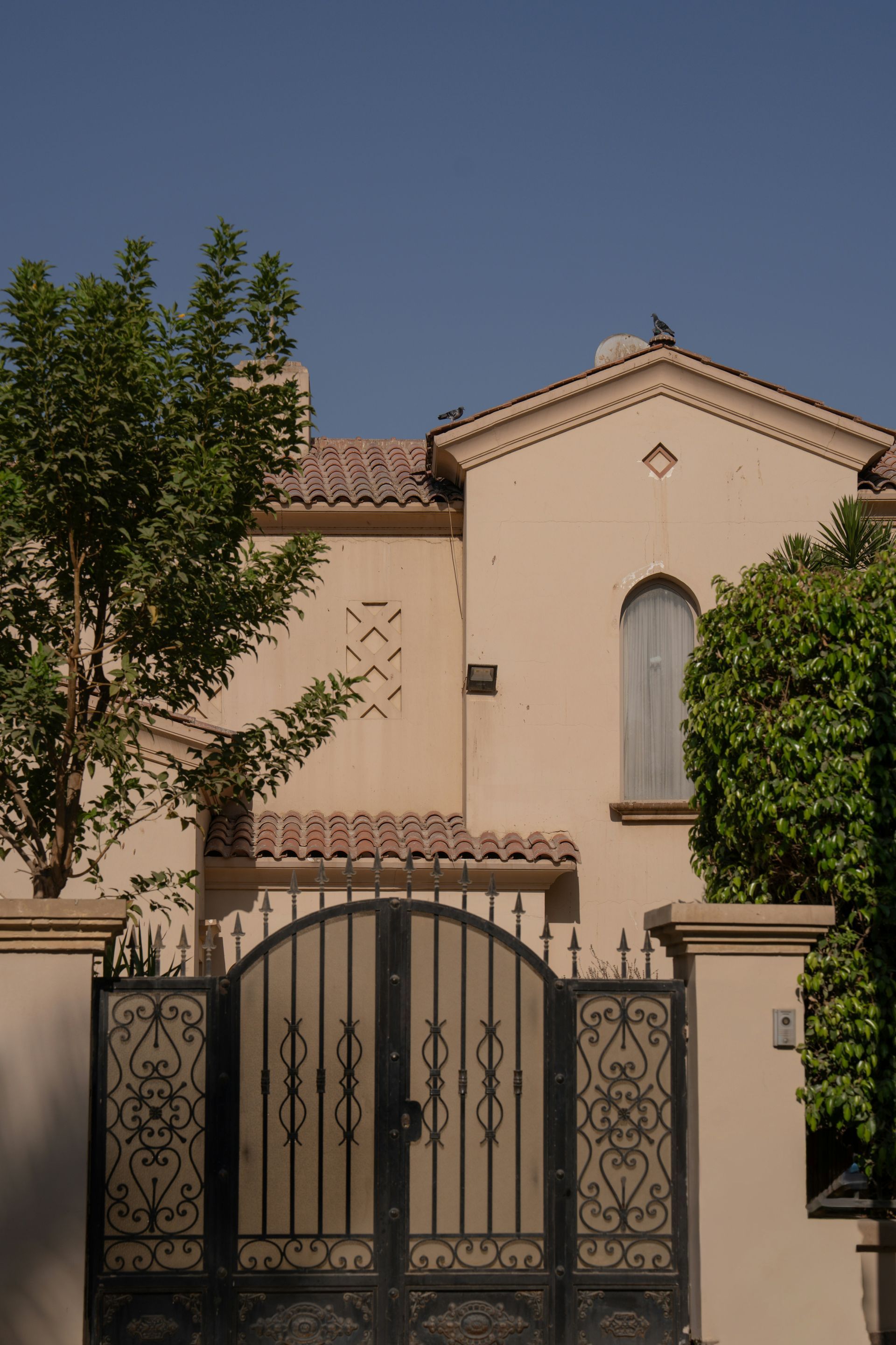 Beige stucco house with tiled roof, arched window, and black wrought-iron gate under a clear blue sky.