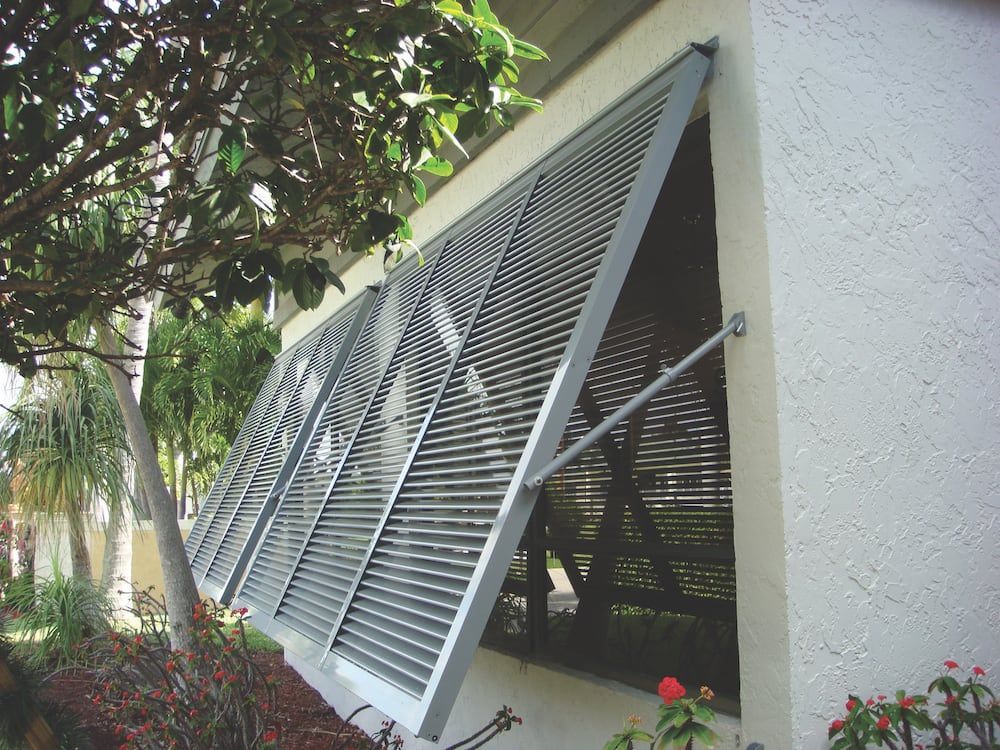 White Bahama shutters open outward on a house exterior beside green foliage.