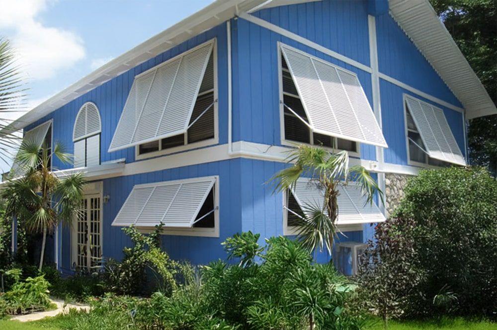 Blue two-story house with white-trimmed windows and Bahama shutters surrounded by lush greenery.