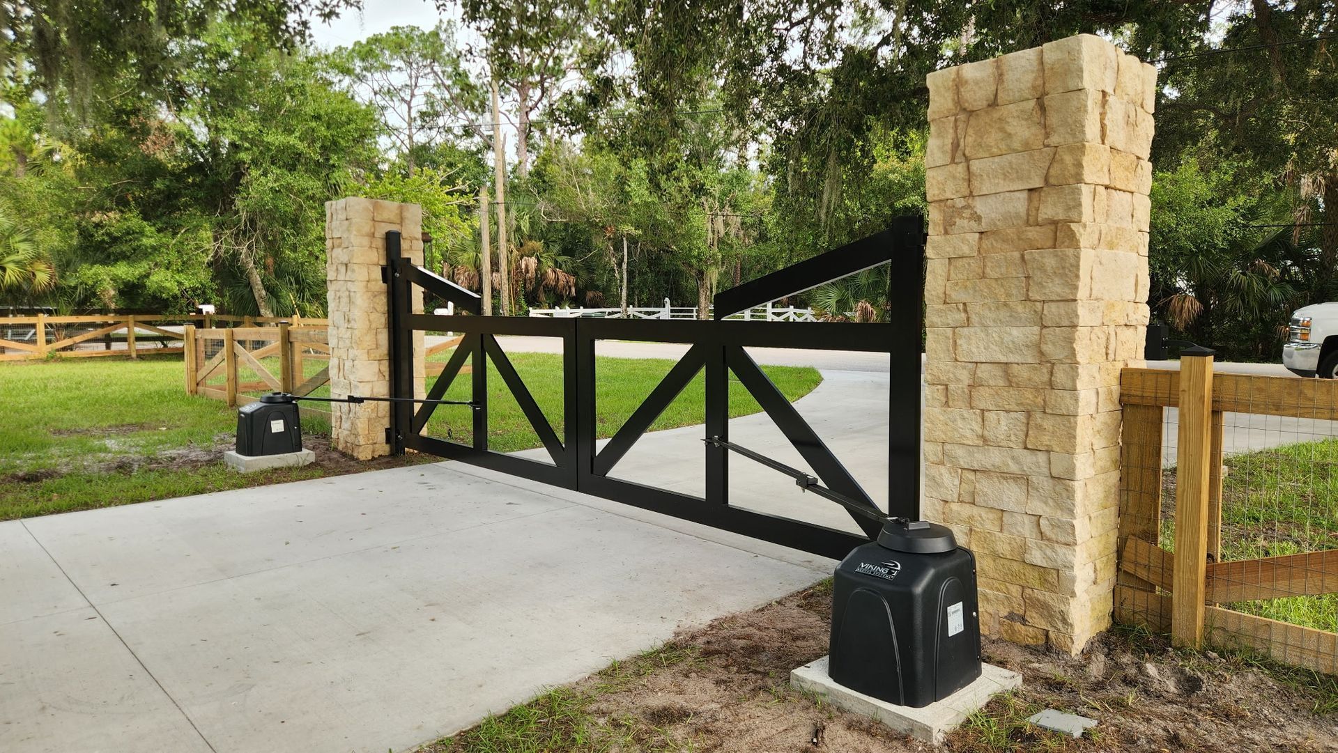 Black wooden gate between stone pillars at a driveway entrance, with trees and greenery beyond.