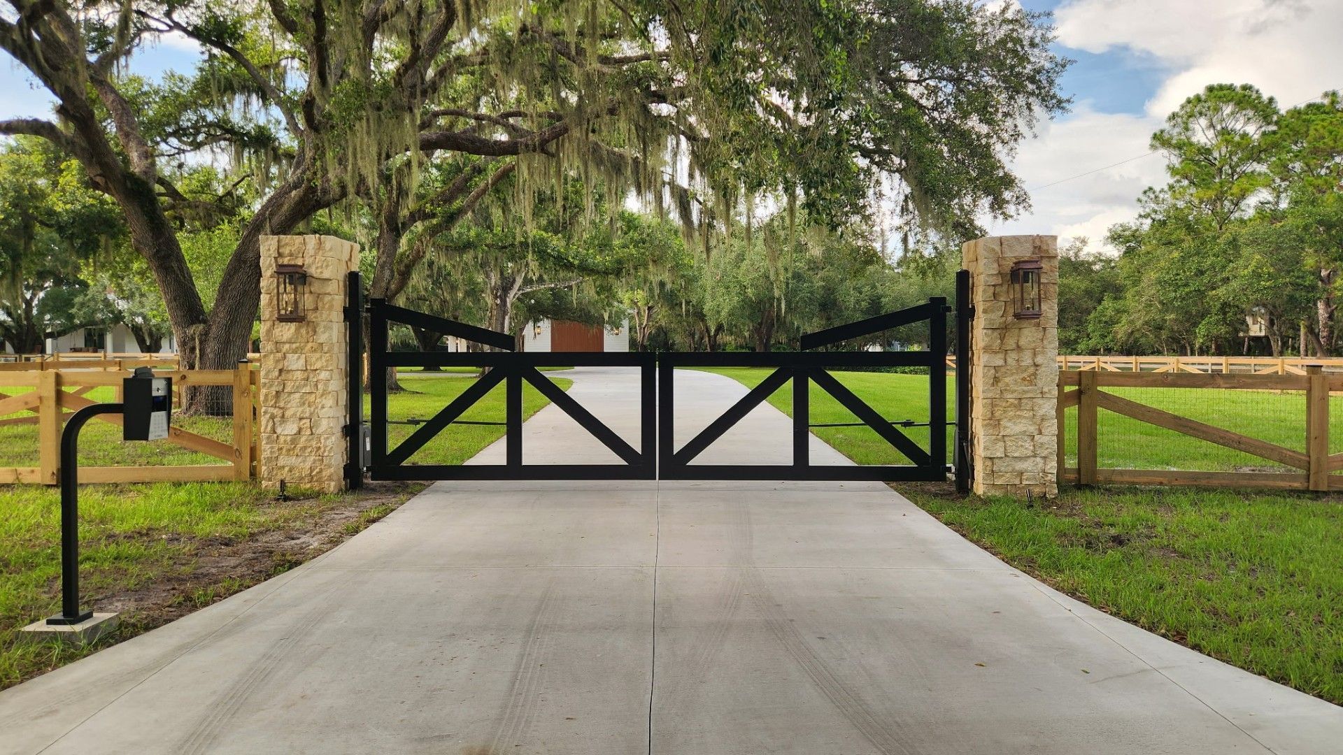 Black metal gate between stone pillars at the end of a concrete driveway, bordered by trees and grass
