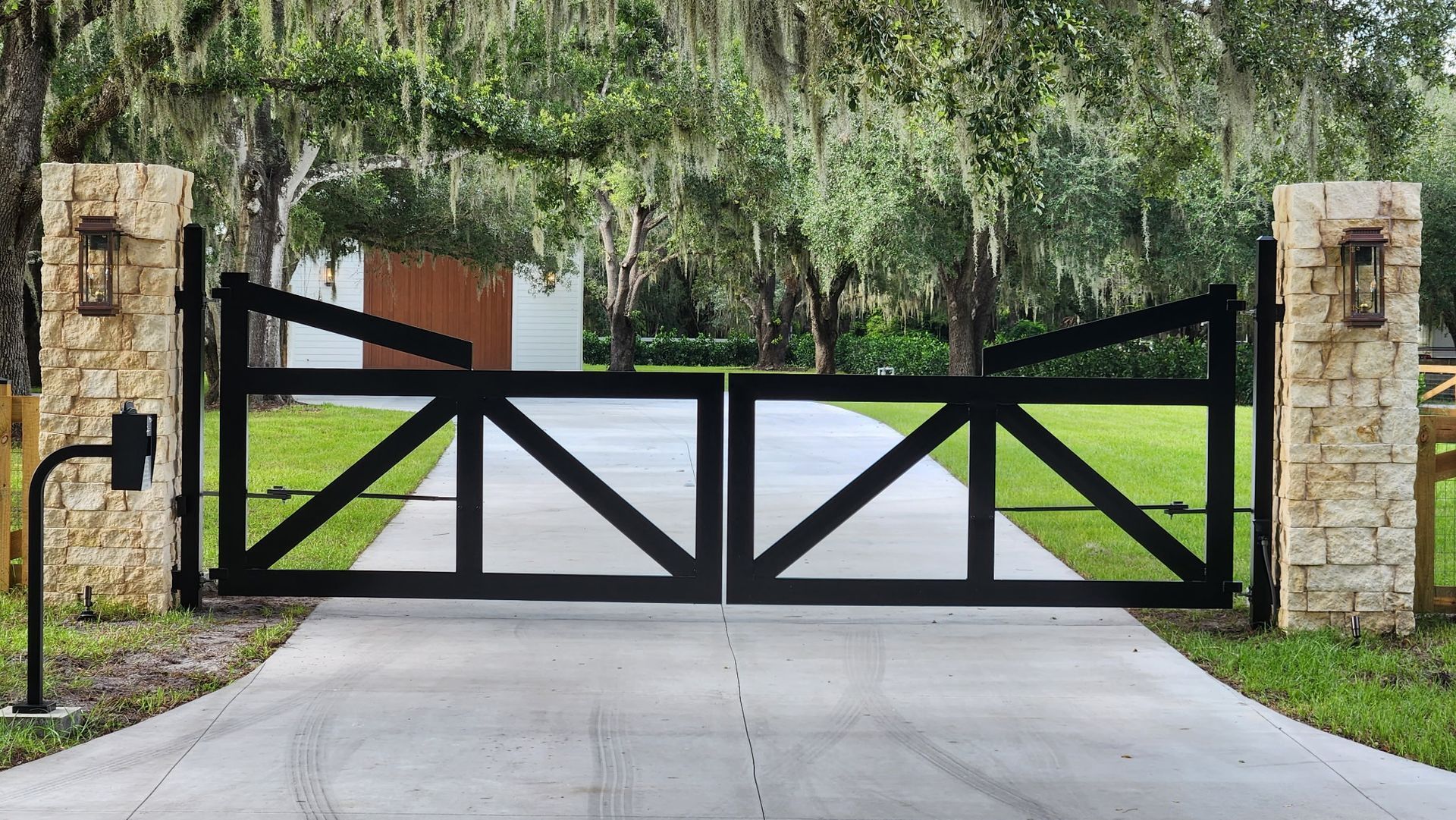 Black wrought-iron driveway gate between stone pillars under hanging trees