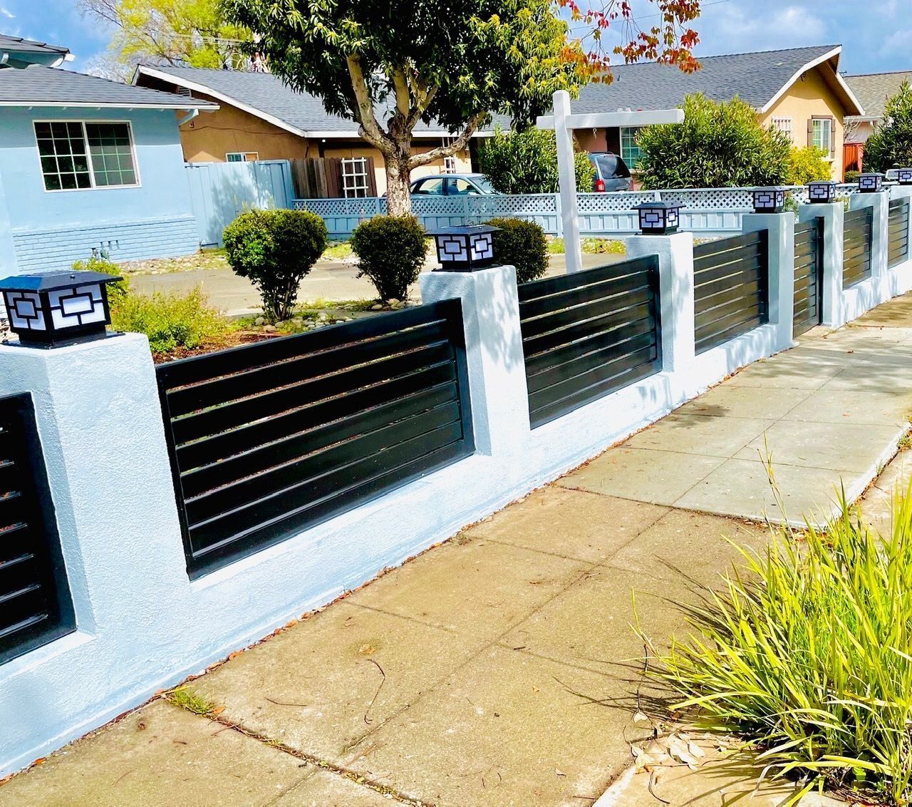 A modern, light blue and black fence with built-in lights lines a sidewalk in front of houses.