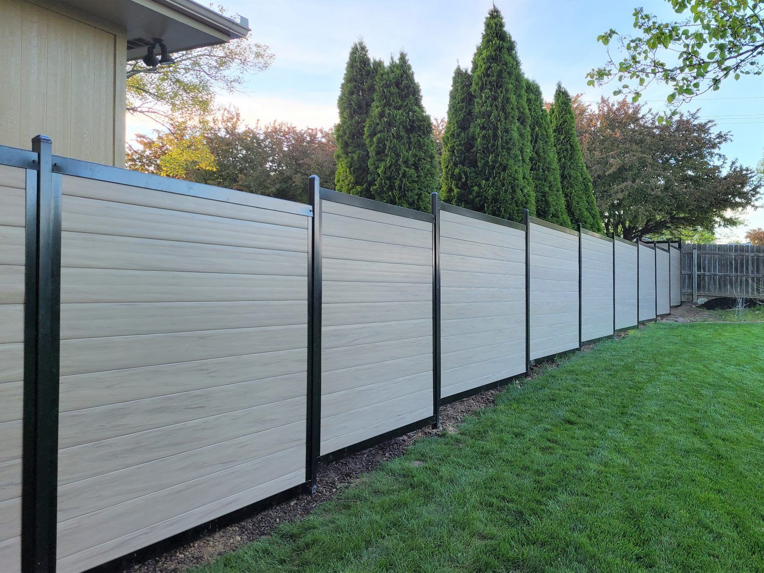 Beige and black privacy fence in a backyard, with green grass and trees.