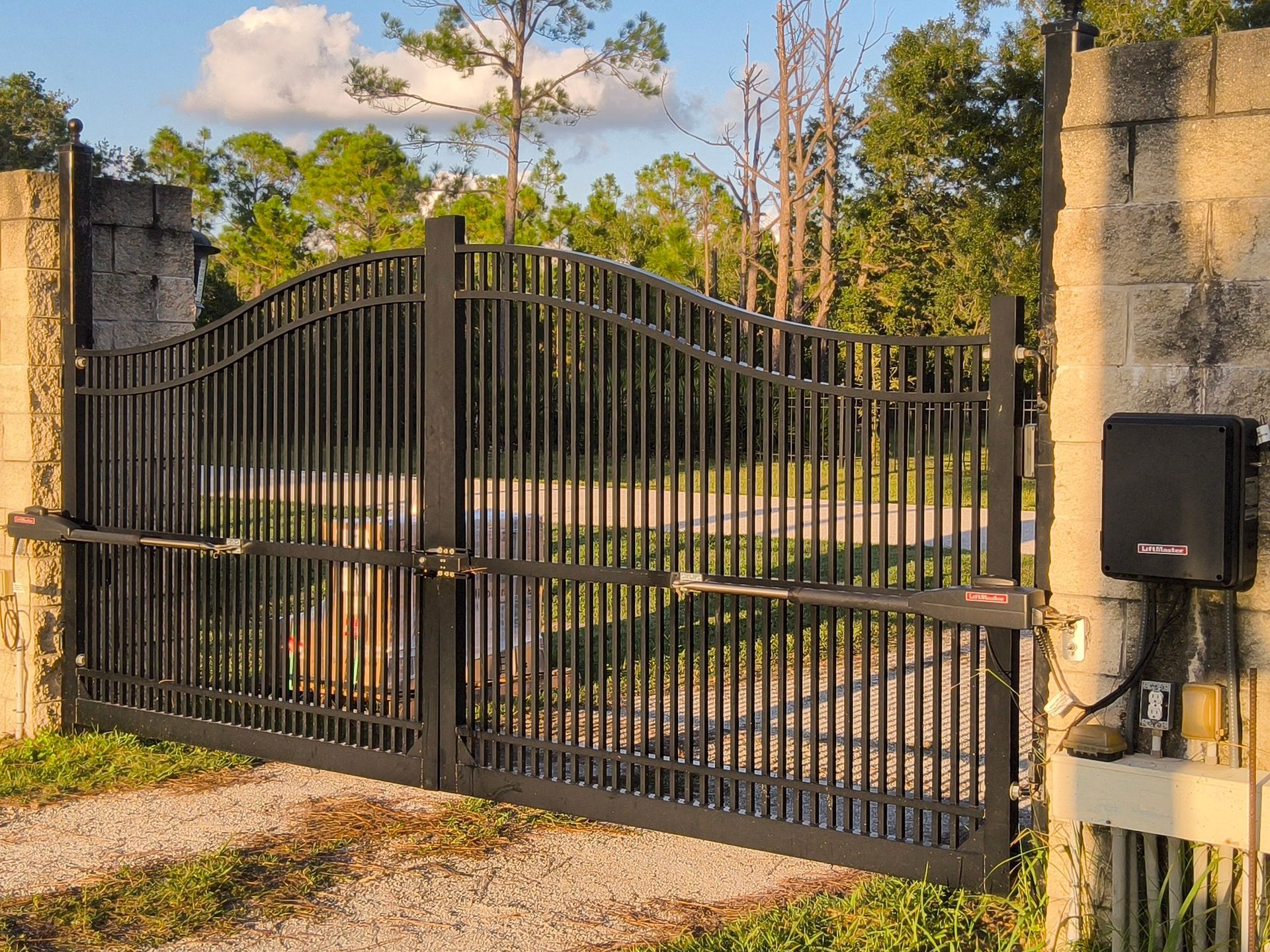 Black metal gate on stone pillars with an automatic gate opener in a driveway.