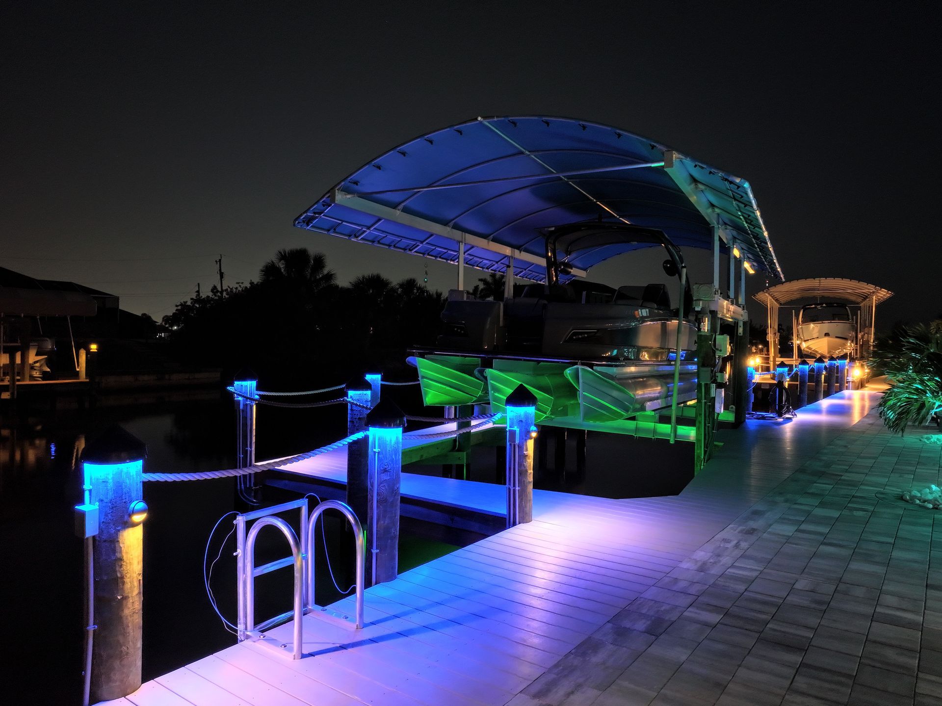 Boat dock illuminated with vibrant blue and green LED lights at night, featuring a covered pontoon boat and glowing pathway