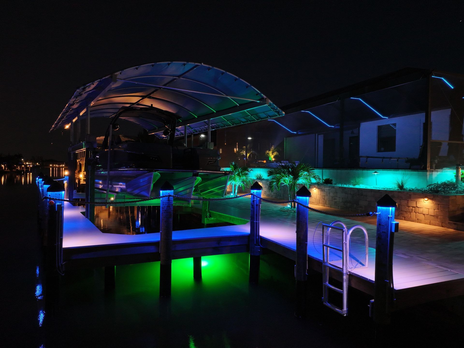 Night view of a waterfront property with illuminated dock, palm trees, and a pool area glowing in blue, green, and amber lights.