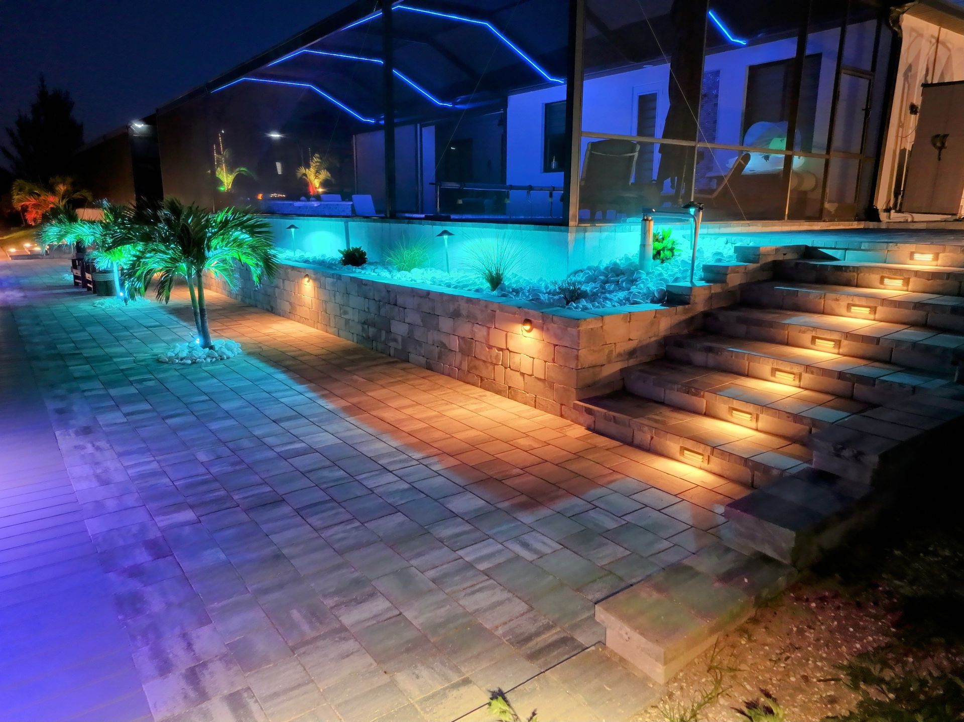 Modern backyard with a pool and patio area, enhanced by colorful landscape lighting, stone steps, and tropical plants at night.