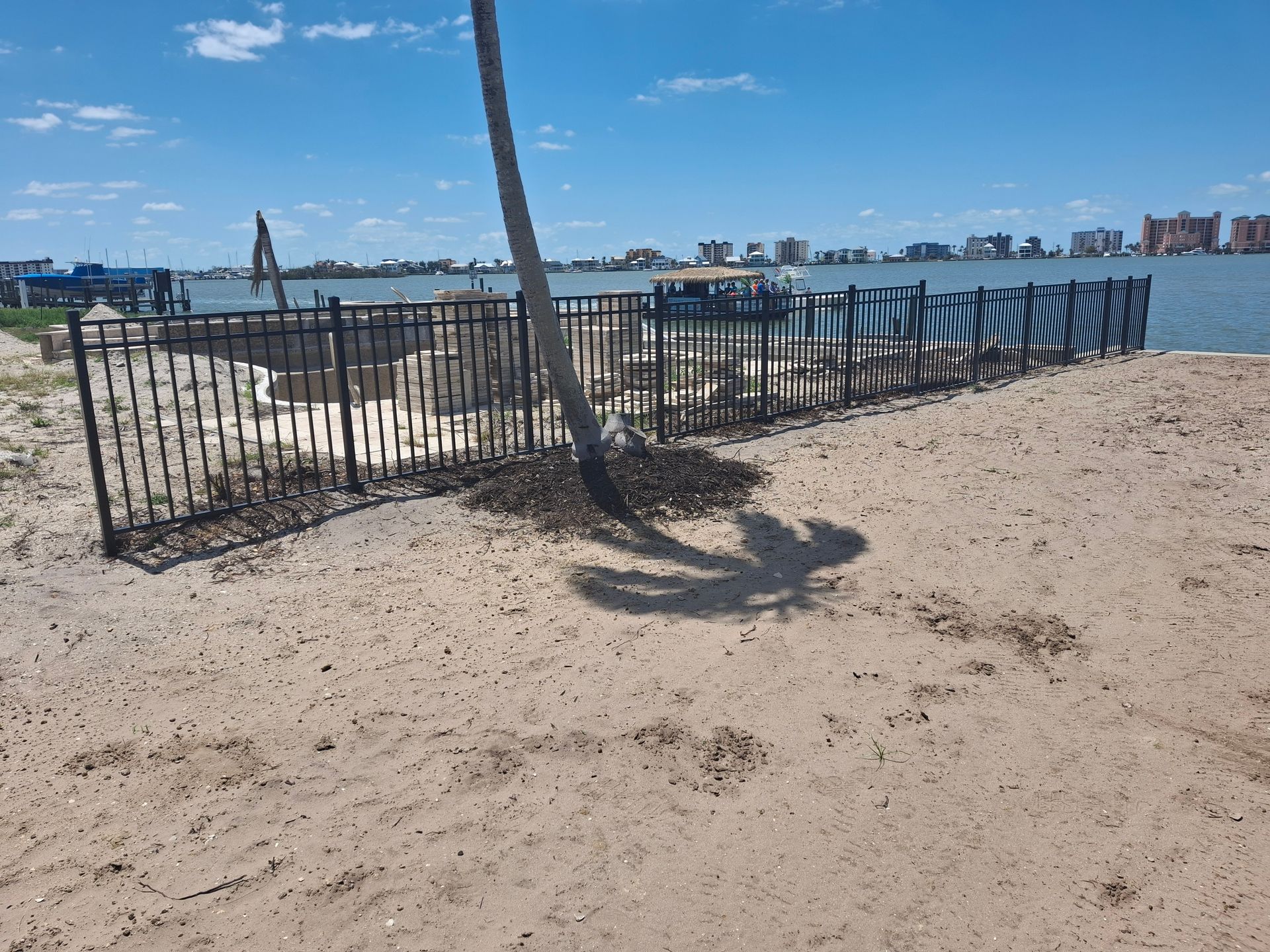 A fence surrounds a sandy beach with a palm tree in the foreground.