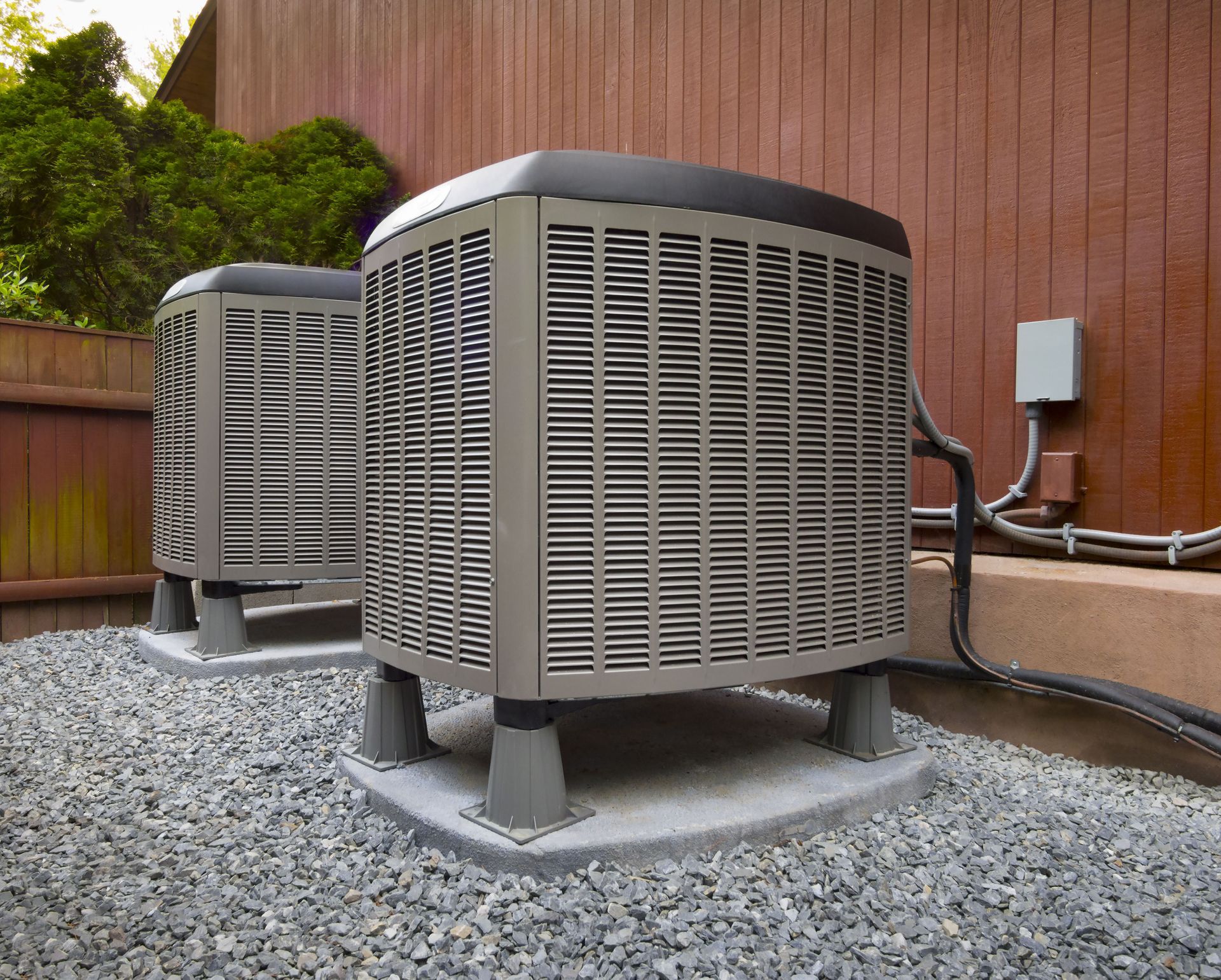 Two outdoor air conditioning units in front of a wooden fence on a bed of gravel.