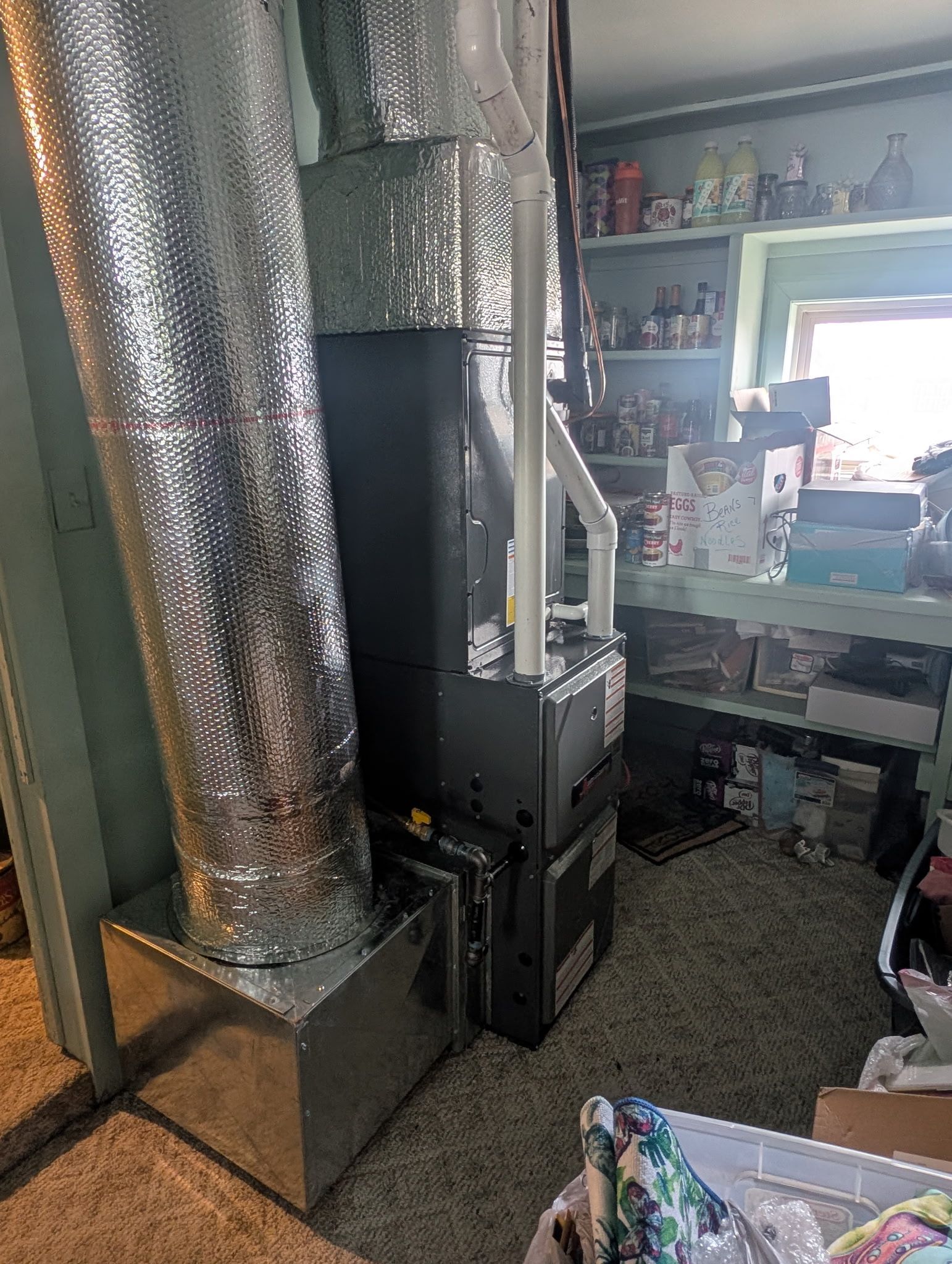 furnace unit with silver ductwork, in a room with shelves and a window