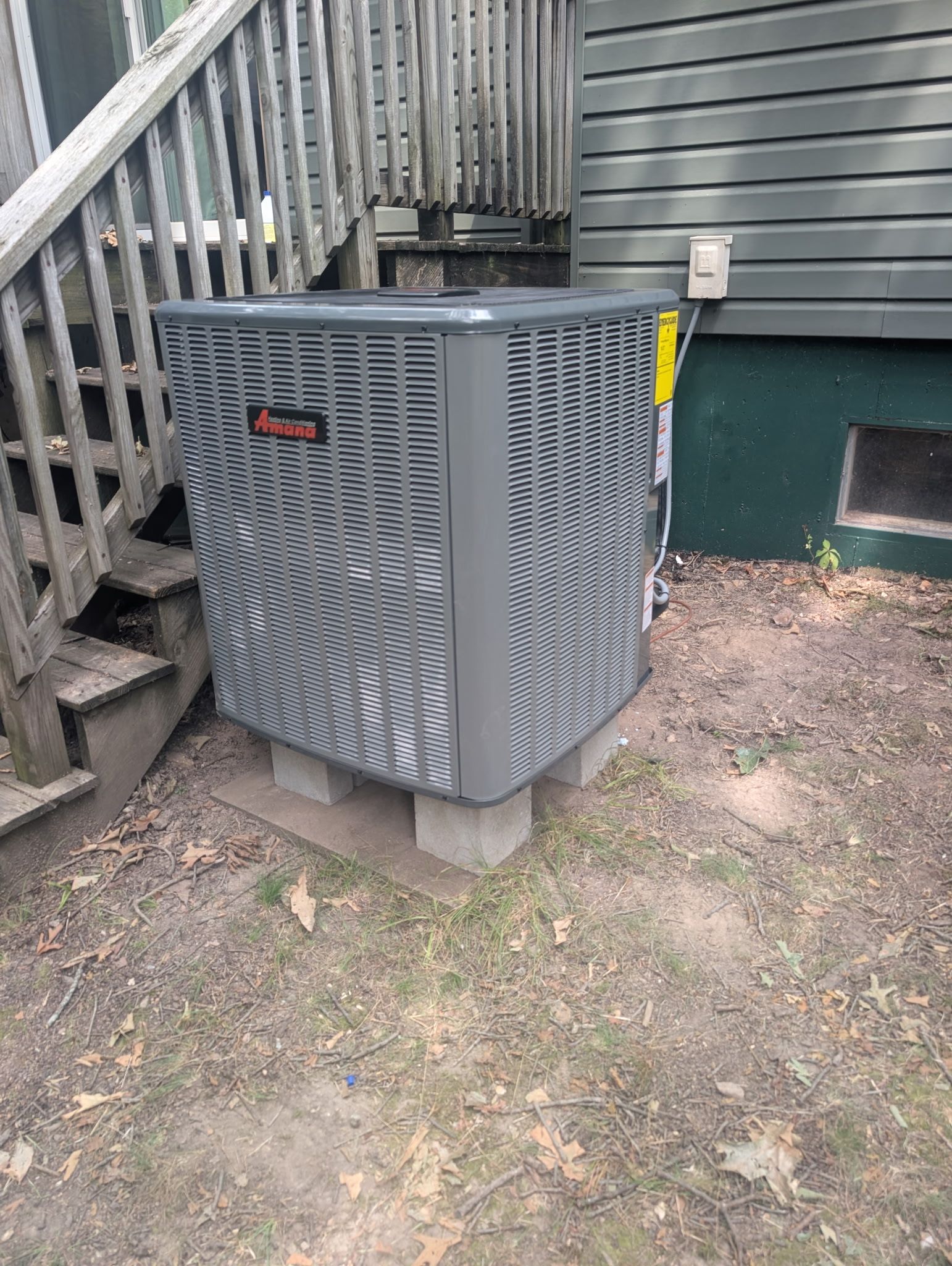 gray Trane air conditioning unit, outdoors next to wooden stairs and a house with green foundation