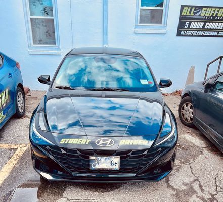 Black Hyundai student driver car parked in front of a building with a blue car next to it.