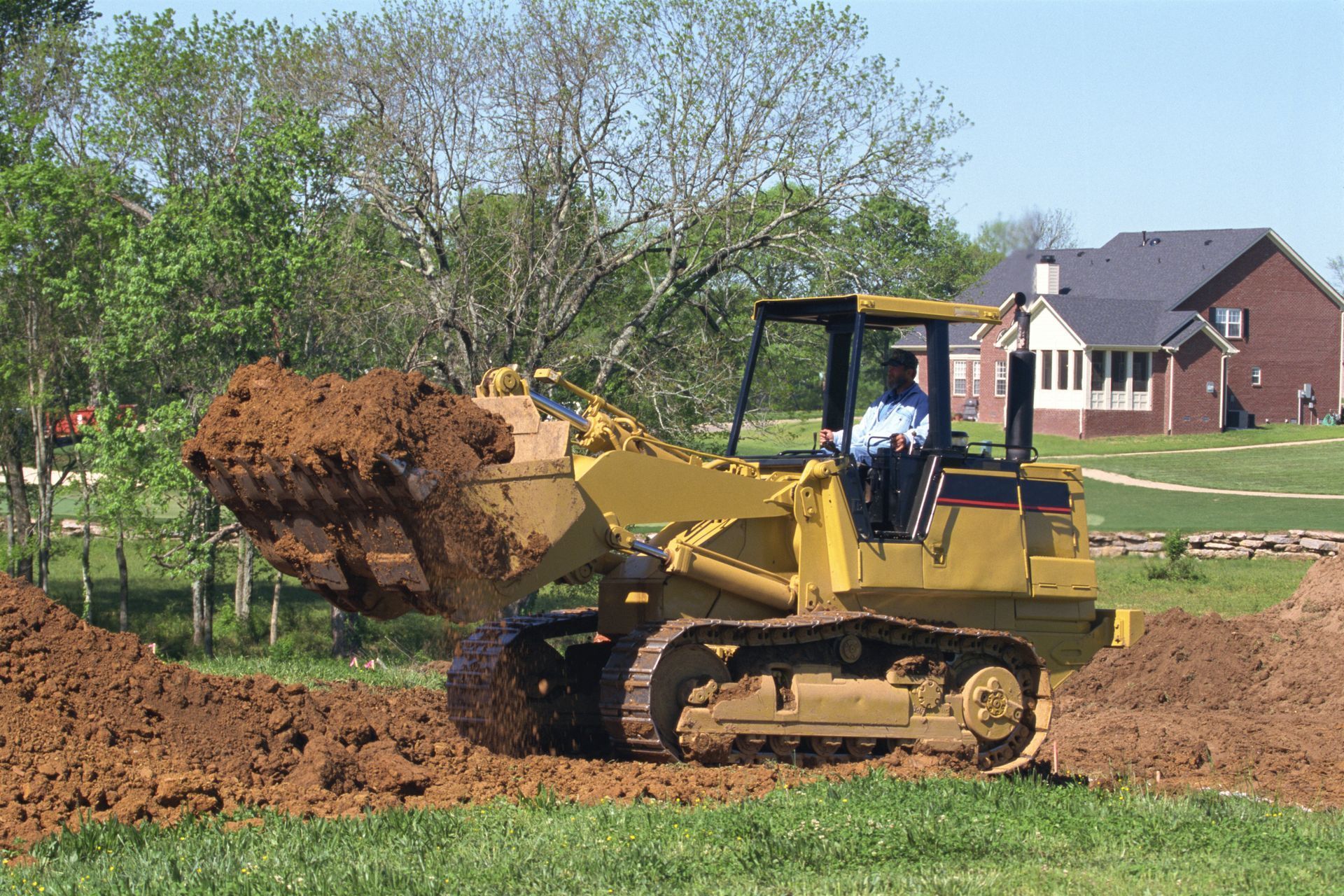 A yellow crawler loader moves a bucket of dirt in an outdoor, grassy residential area with a house in the background.