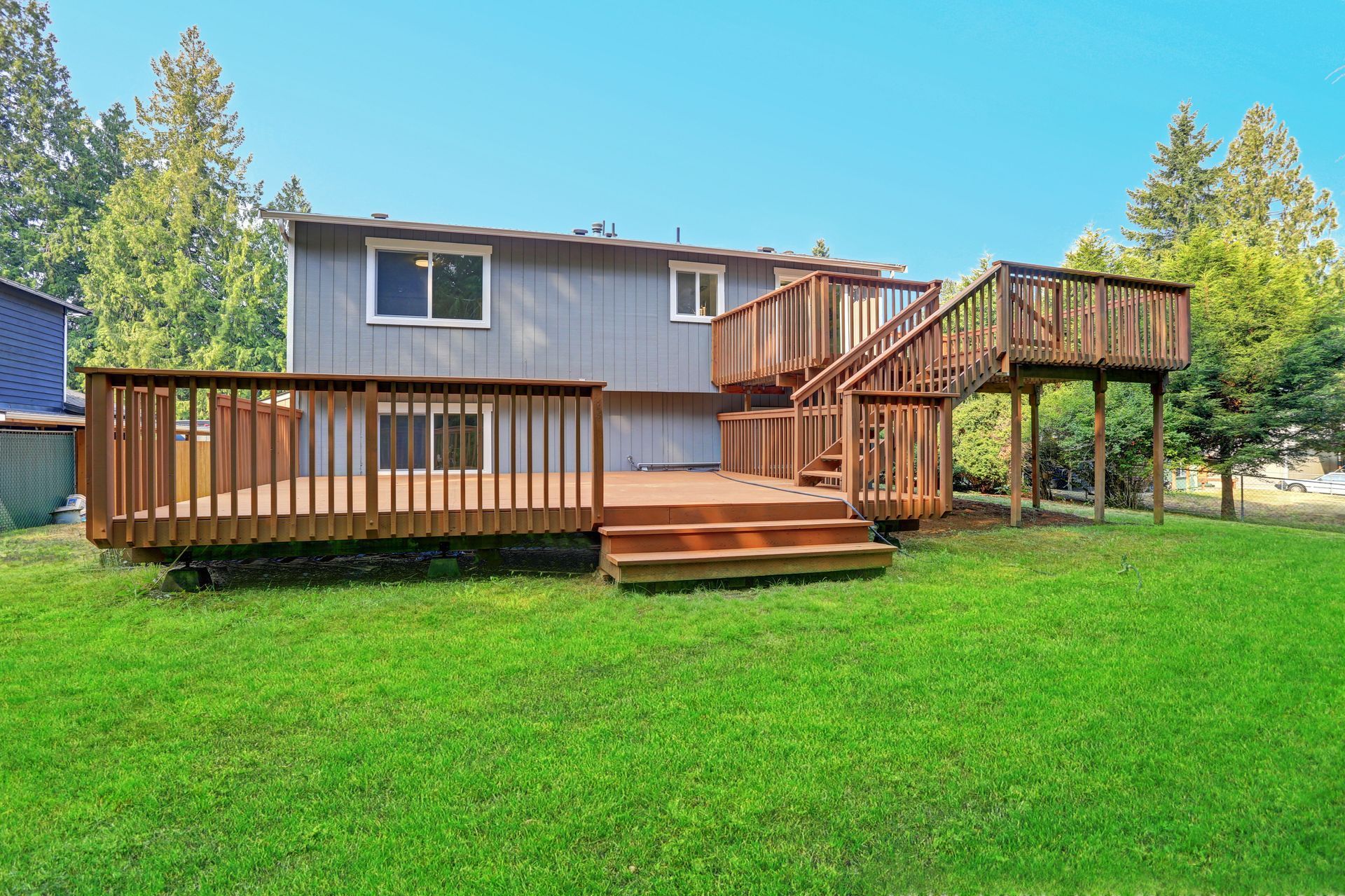 A two-story grey house features a large, multi-level wooden deck with stairs in a grassy backyard under a clear blue sky.