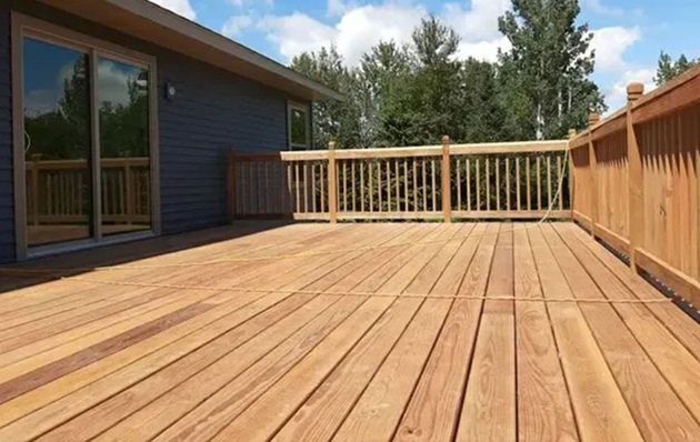 A wooden deck attached to a blue house with trees in the background under a blue, cloudy sky.