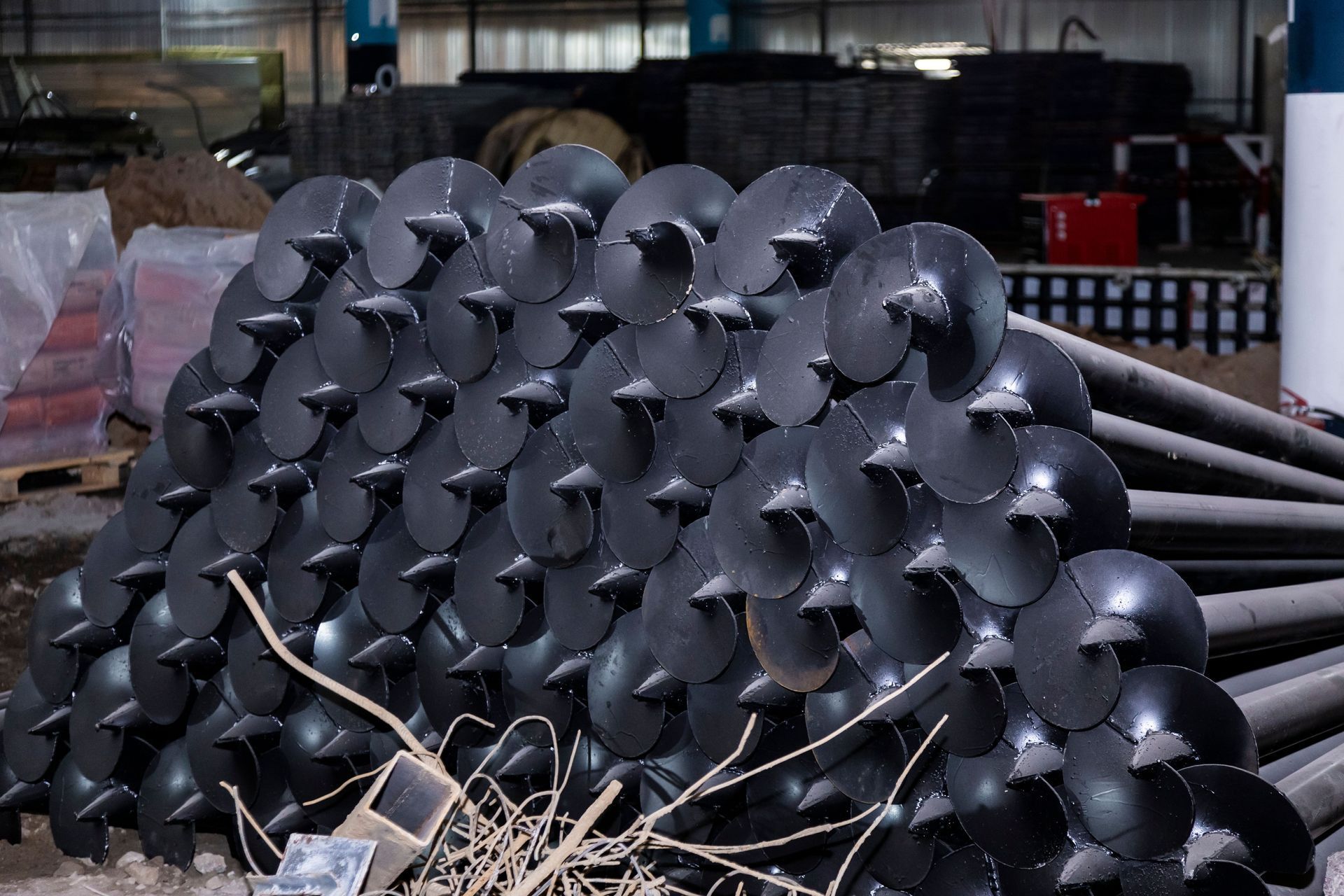 A large pile of dark gray helical pile foundations stacked horizontally in a warehouse.