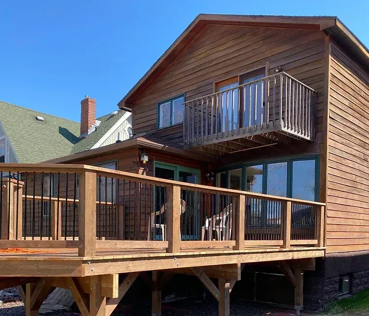 A wooden two-story house with a large ground-level deck, a second-story balcony, and a green-roofed neighbor house nearby.
