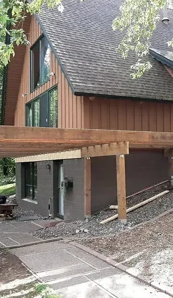 A two-story wood-sided house with a dark brown lower level, a shingled roof, and an unfinished wooden patio frame.
