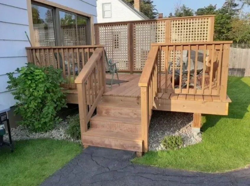 A wooden deck with a staircase and lattice privacy screen attached to the side of a white house, set in a grassy yard.