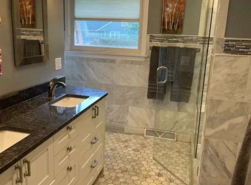 A bathroom with a white vanity, dark stone countertop, marble tiled walls, a glass-enclosed shower, and hexagonal floor.
