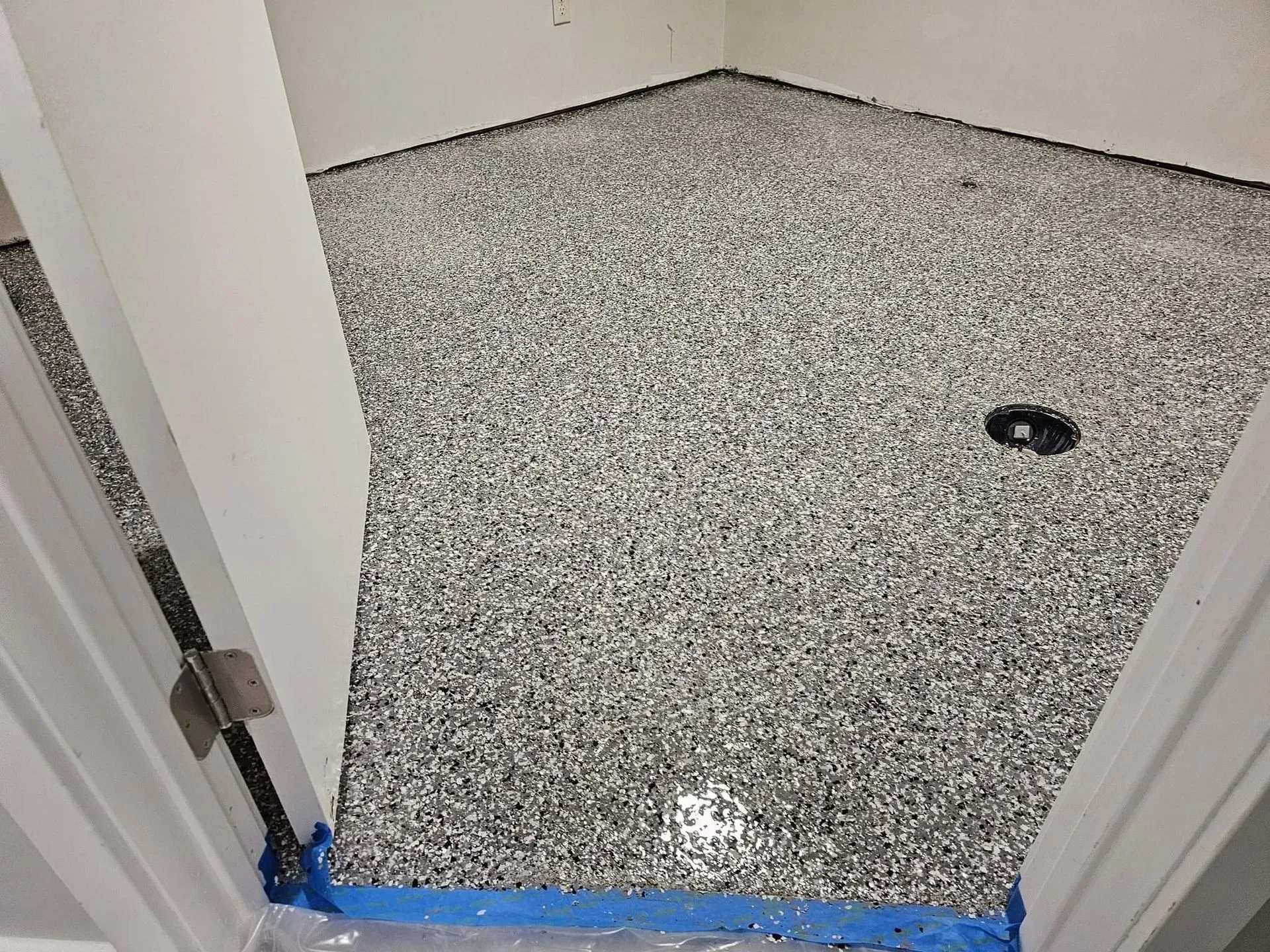 A room with a newly applied gray and black speckled epoxy floor coating, viewed through an open doorway with blue tape.