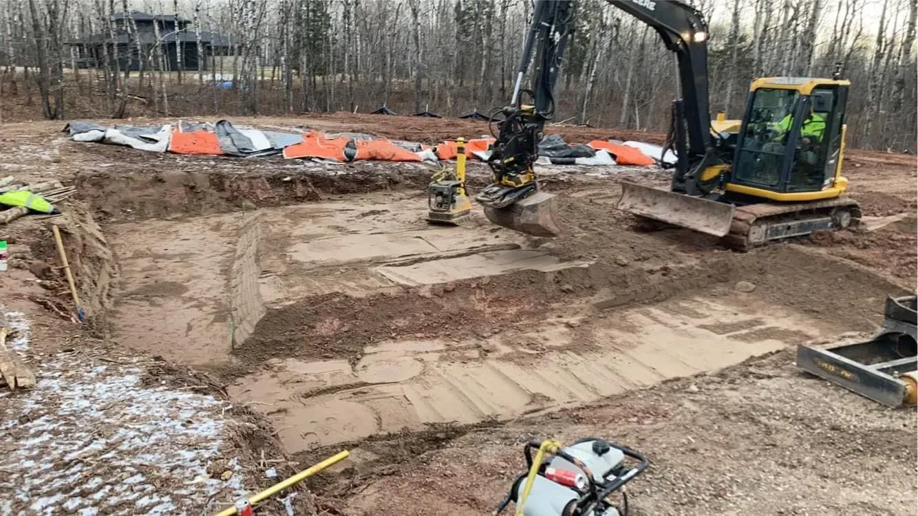 A yellow excavator digs a square foundation trench in a muddy construction site outdoors.