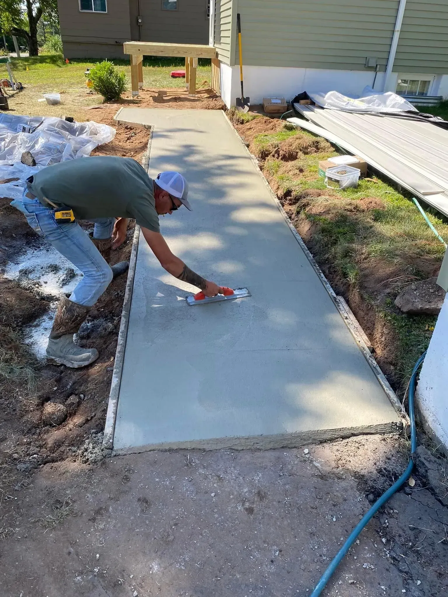 A person in a green shirt and white cap smoothing wet concrete on a new walkway path next to a house.