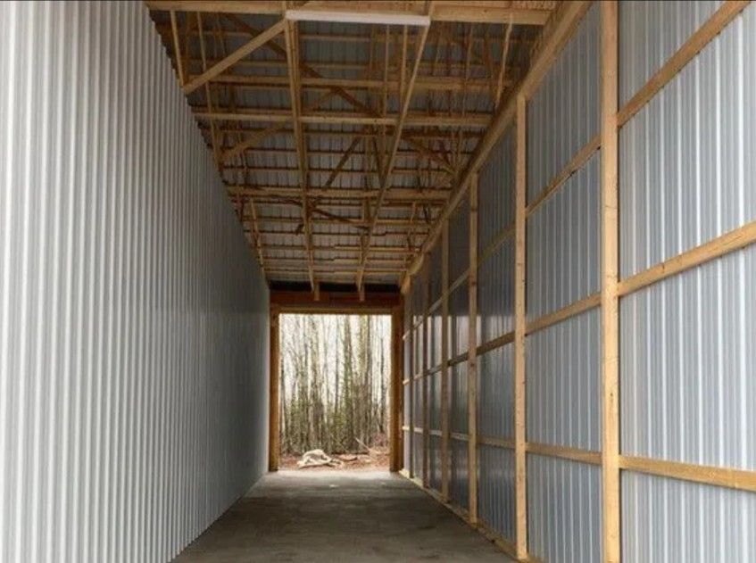 A hallway inside a metal-walled building with a wooden truss ceiling, looking out toward a forest through an open doorway.