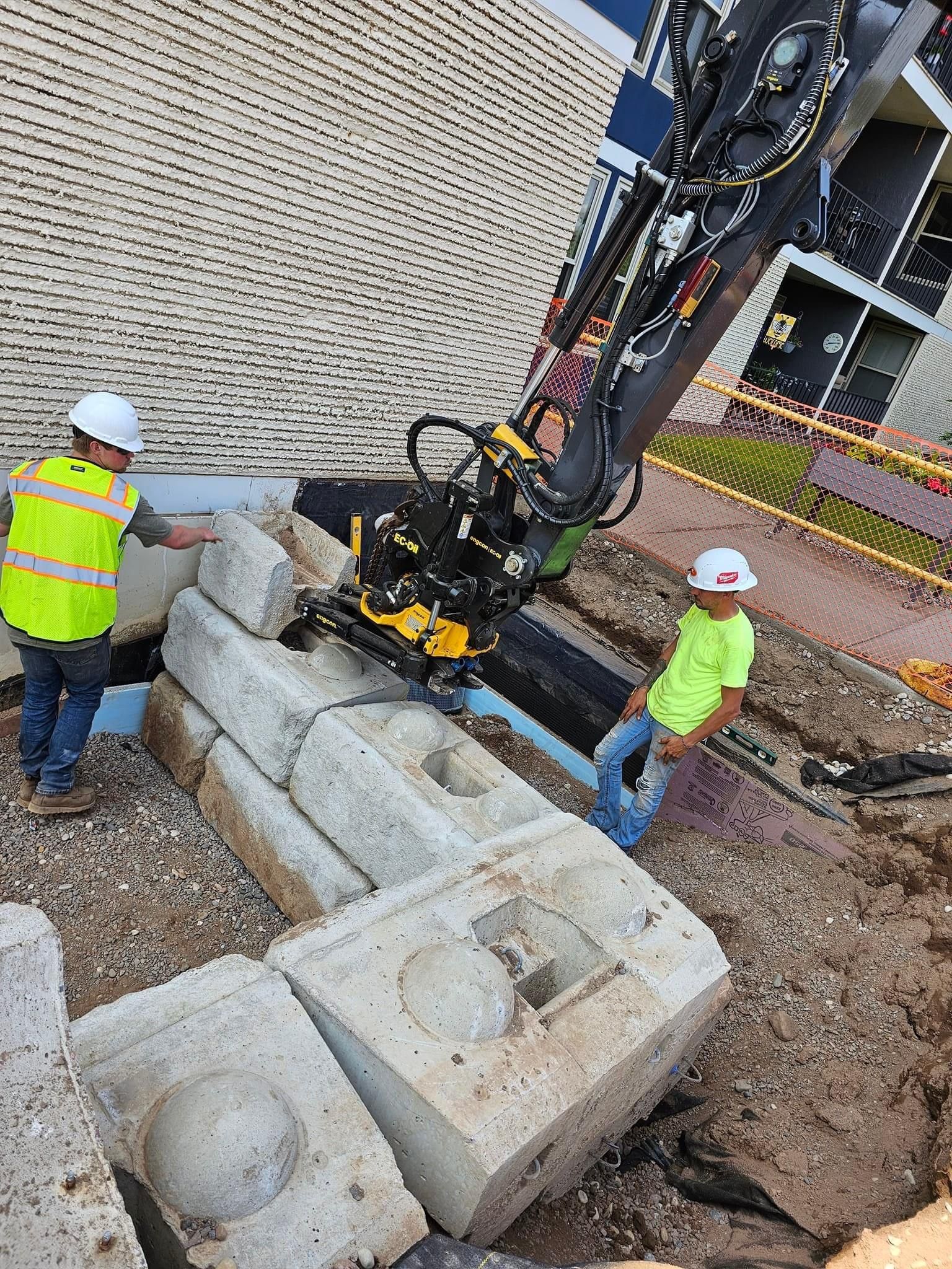 Two construction workers in high-visibility vests and hard hats watch an excavator place large concrete blocks.