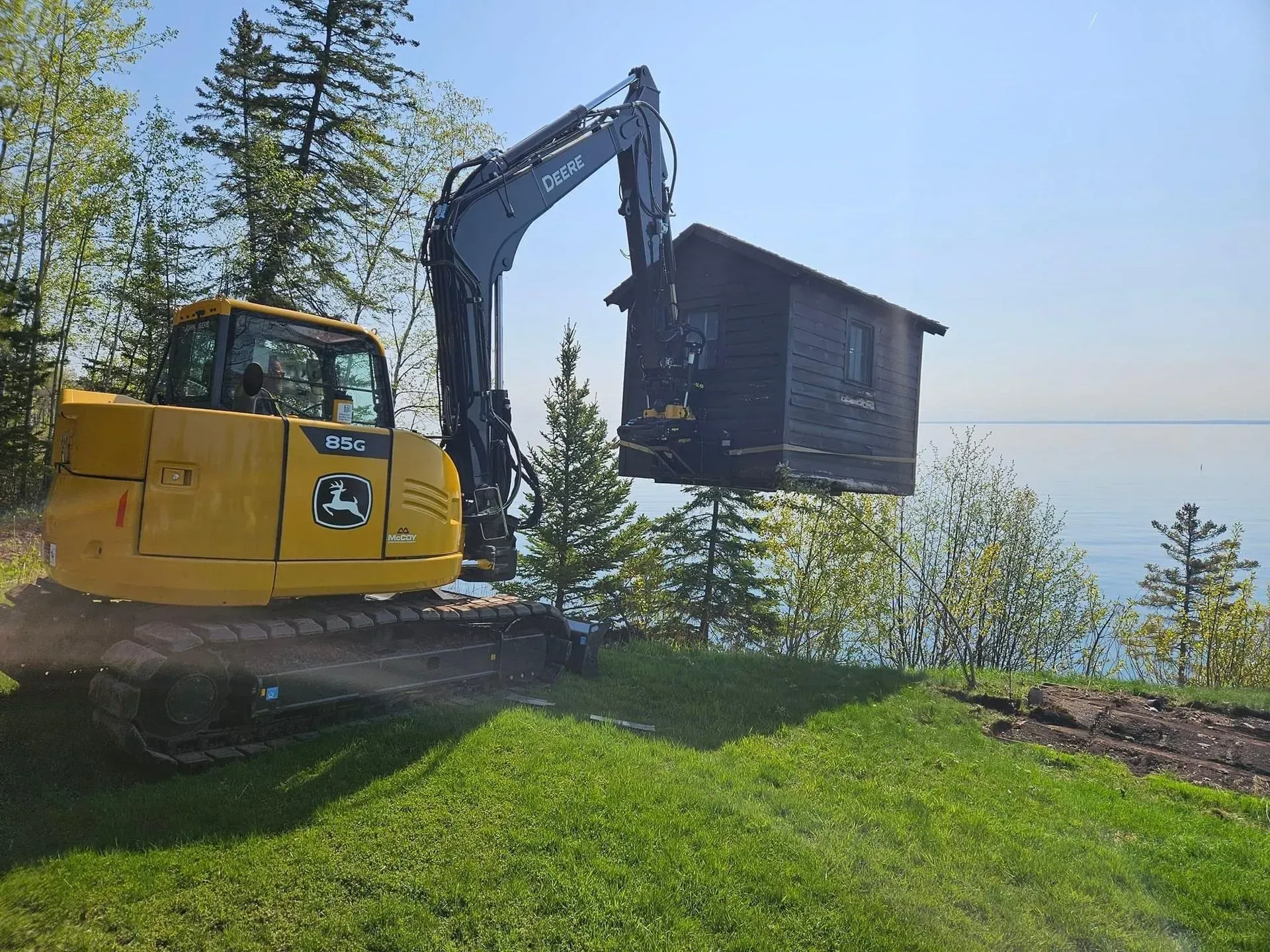A yellow John Deere excavator lifting a small wooden shed on a grassy hill overlooking a body of water.