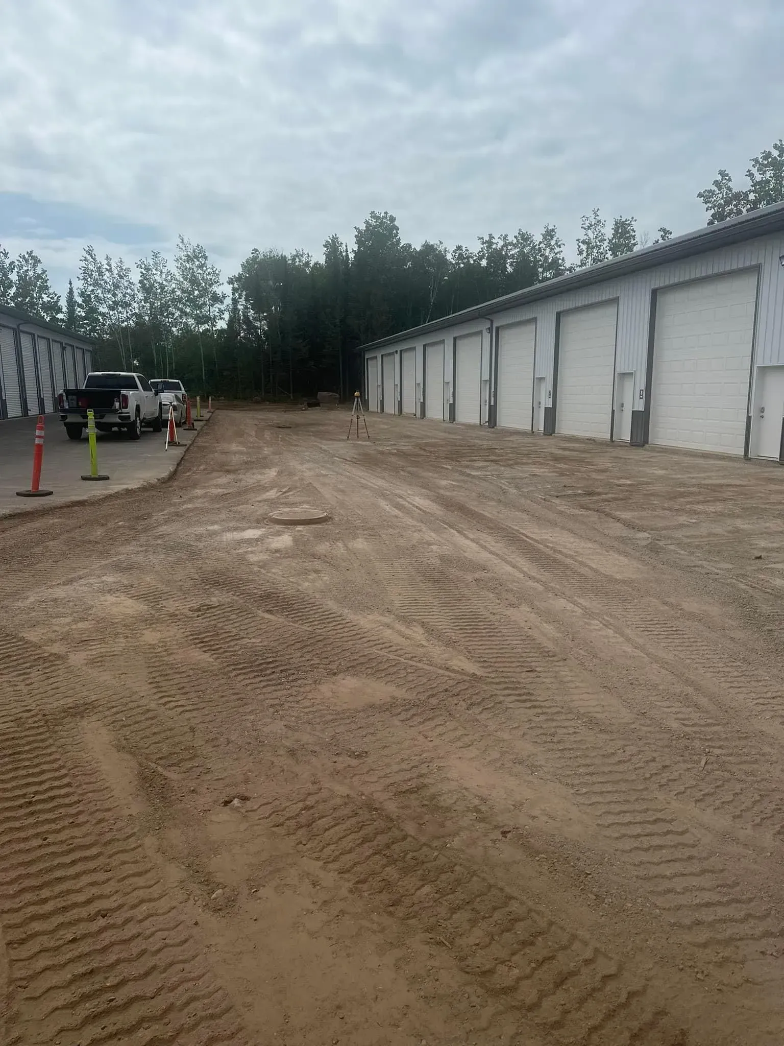 A gravel construction site featuring a row of white storage units with rolling doors, beside a line of trees.