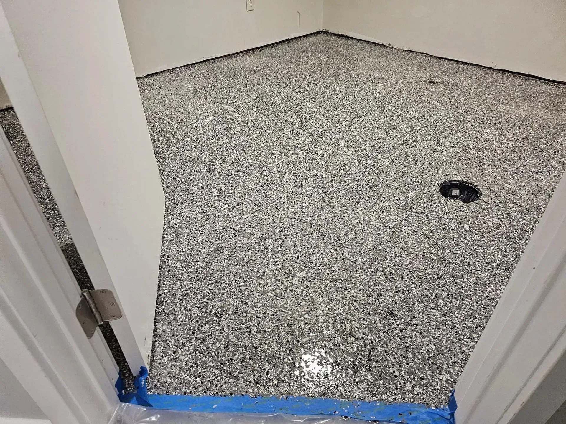 A room floor covered in a grey, black, and white speckled epoxy coating, viewed through a doorway with blue painter's tape.