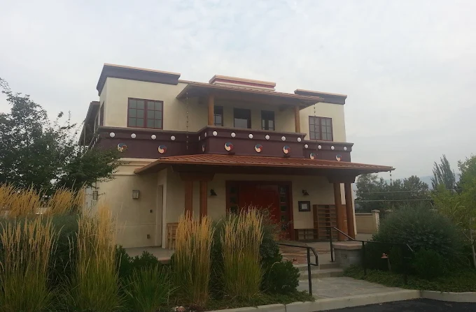 Two-story building with tan stucco walls, brown trim, and a terracotta roof. Tall grasses stand in front.