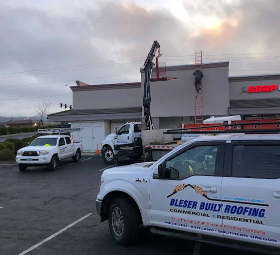 Construction workers on the roof with trucks and a lift. Bleser Built Roofing logo on truck. Building exterior.