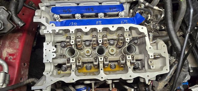 Engine cylinder head and valve train exposed in a car engine bay, with a blue cover at the top.