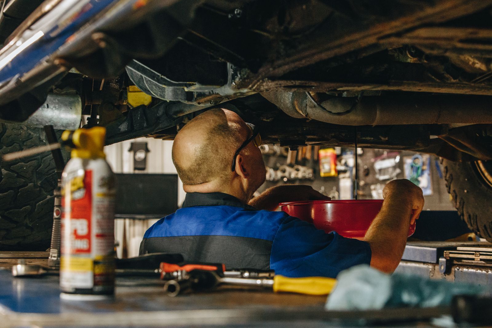 Mechanic working under a car in a garage, reaching toward the underside near tools and a fire extinguisher.