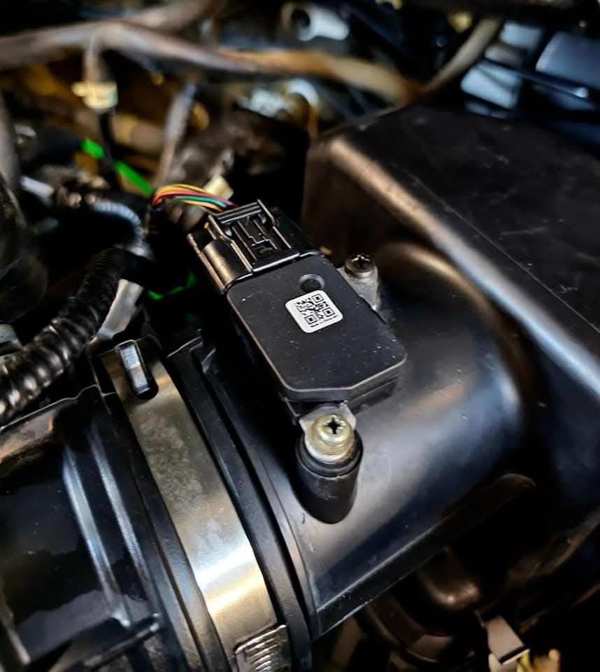 Close-up of a car engine sensor mounted on a black intake component with wiring harness attached